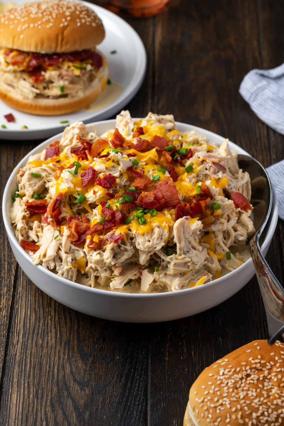 A bowl of Instant Pot crack chicken topped with crumbled bacon, cheese, and chives with a crack chicken sandwich on a plate in the background.