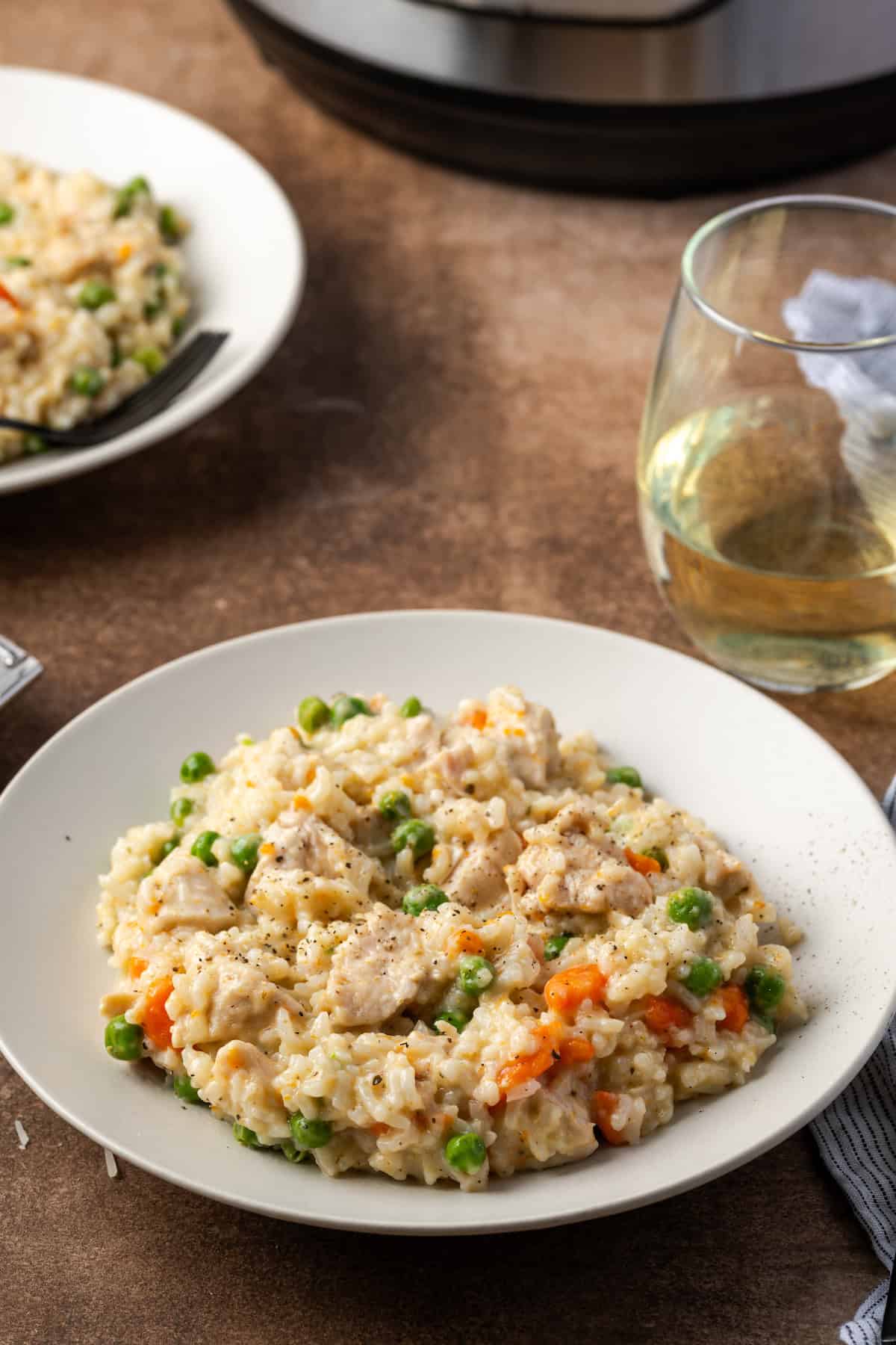 A serving of instant pot chicken and rice in a white bowl next to a stemless glass of white wine, with the corner of the Instant Pot in the background.