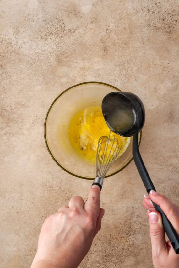 One hand adding a ladleful of hot broth to a bowl of egg yolks, while the other hand uses a whisk to stir and temper the eggs.