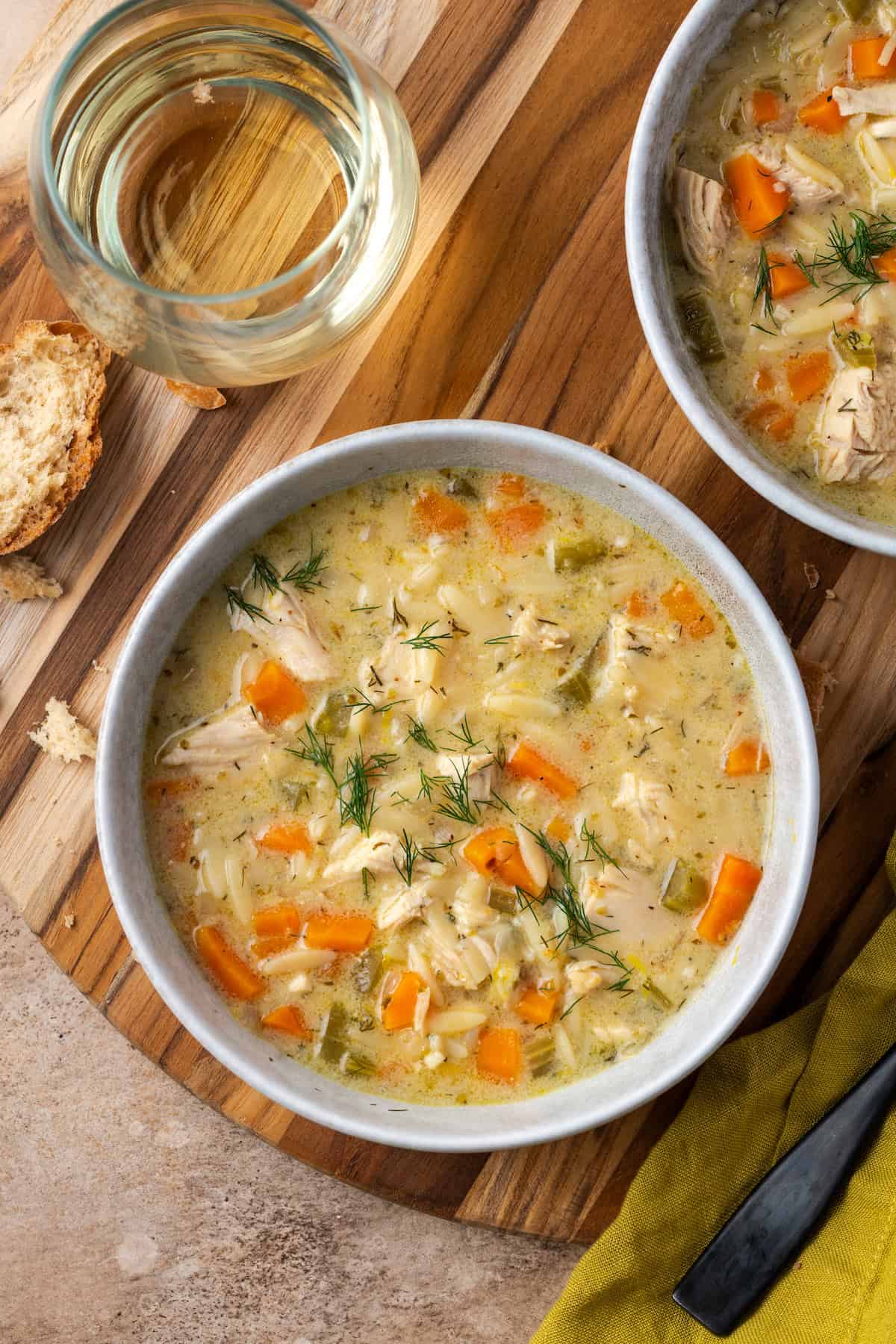 Overhead view of a bowl of lemon chicken orzo soup next to a stemless glass of white wine and a second bowl of soup.
