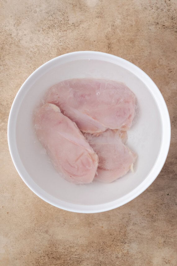 Three chicken breasts brining in salt water in a large white bowl.