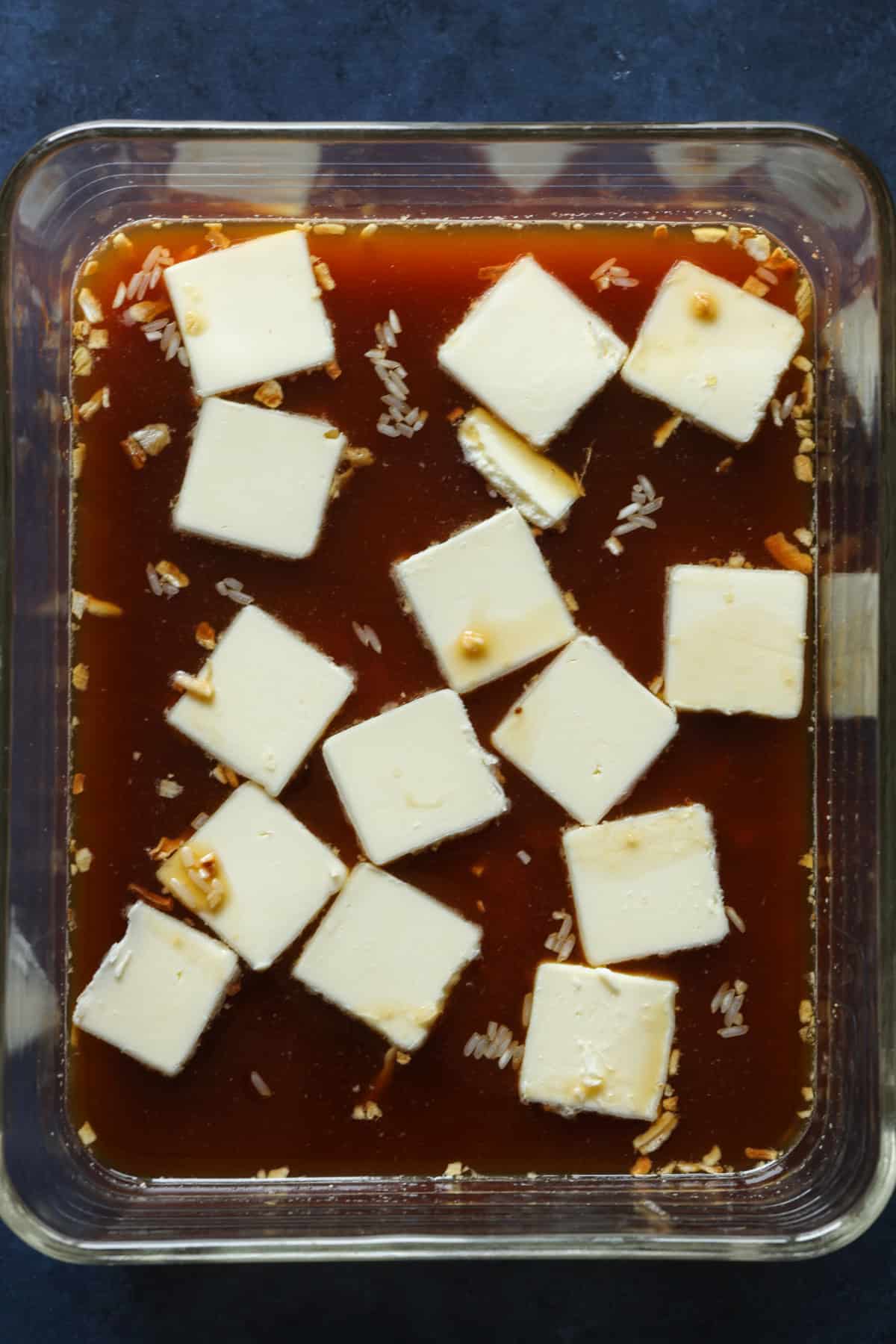 Uncooked rice, beef broth, and onion soup mix in a glass dish topped with slices of butter to be baked.