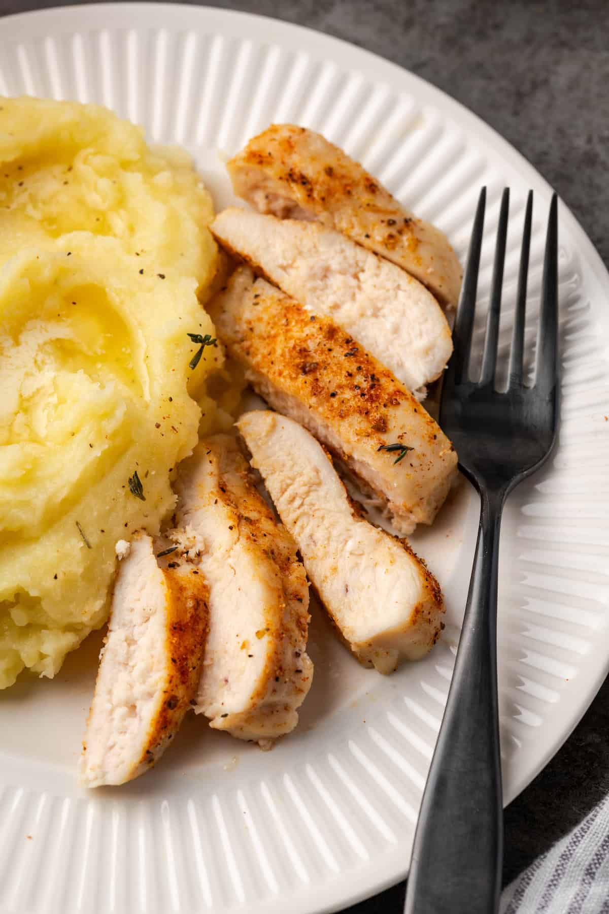 Baked chicken breast cut into slices next to a serving of mashed potatoes and a fork on a white plate.