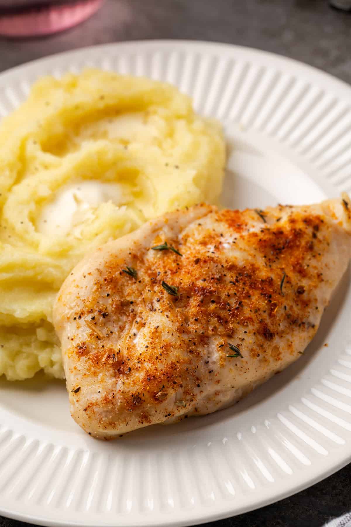 Baked chicken breast next to a serving of mashed potatoes on a white plate.