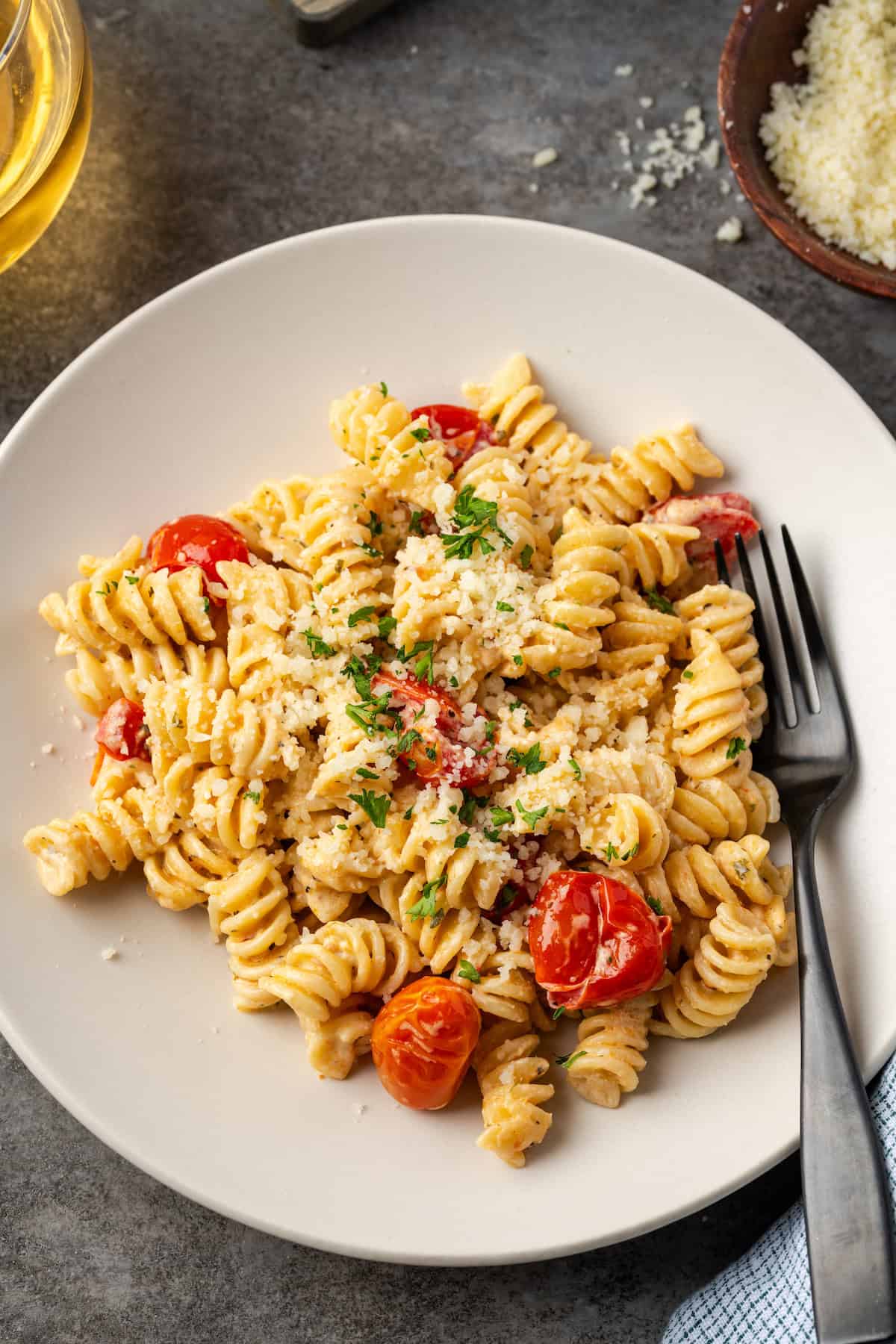 Overhead view of a fork resting on a plate of pasta with Boursin cheese, next to a small plate of parmesan and a glass of wine.