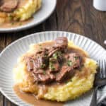 No peek beef tips and gravy served over mashed potatoes on a white plate next to a fork, with a second plate of beef tips in the background.