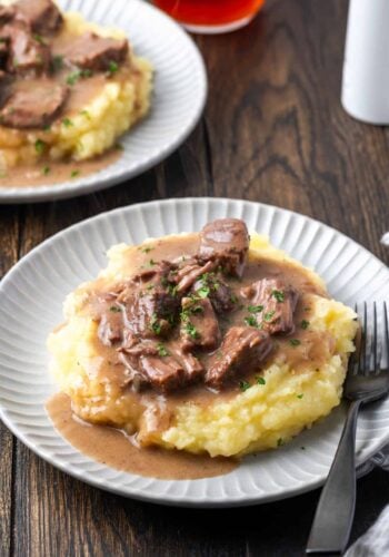 No peek beef tips and gravy served over mashed potatoes on a white plate next to a fork, with a second plate of beef tips in the background.