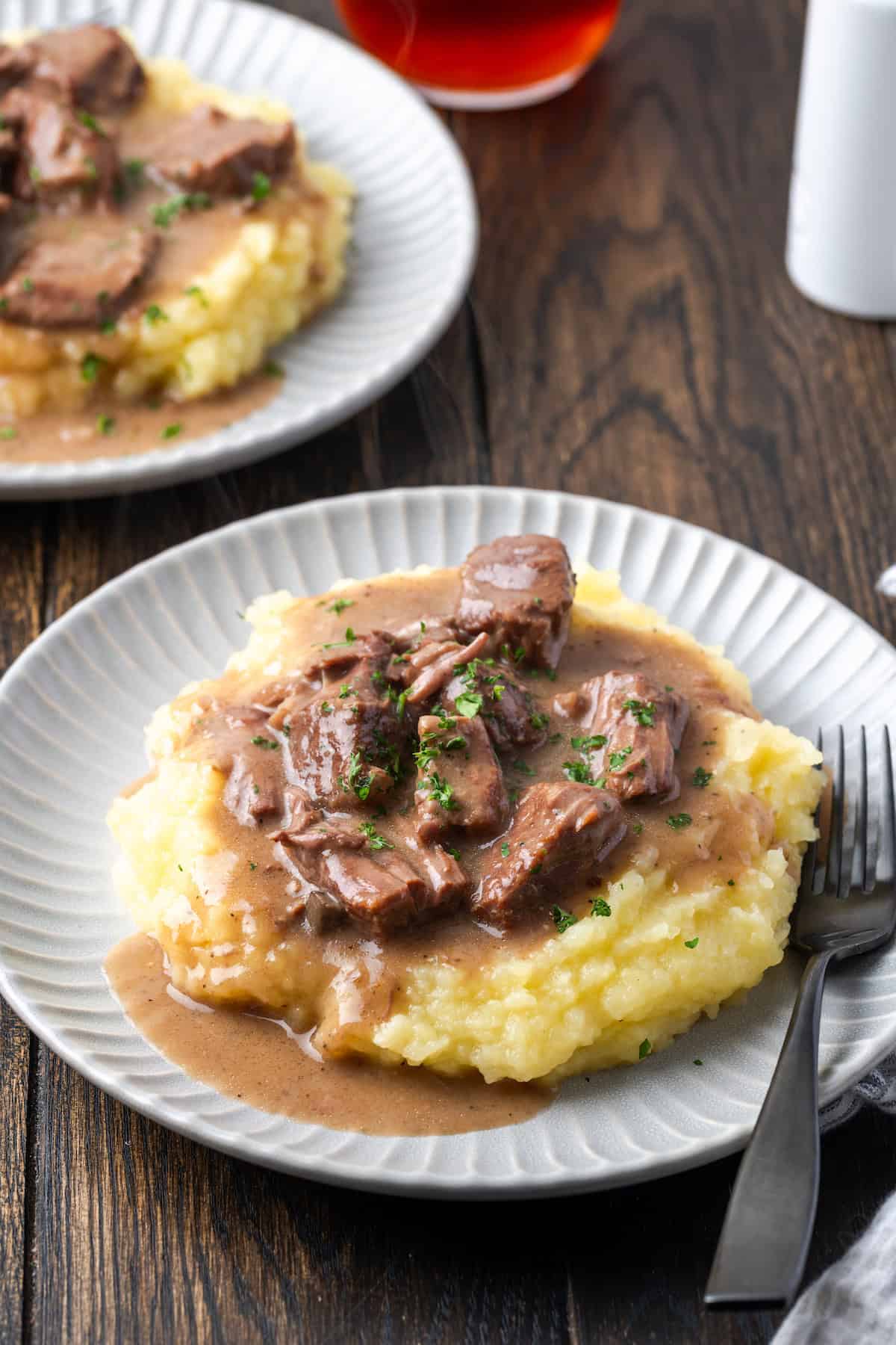 No peeping beef tips and gravy served over mashed potatoes on a white plate next to a fork with a second plate of beef tips in the background.