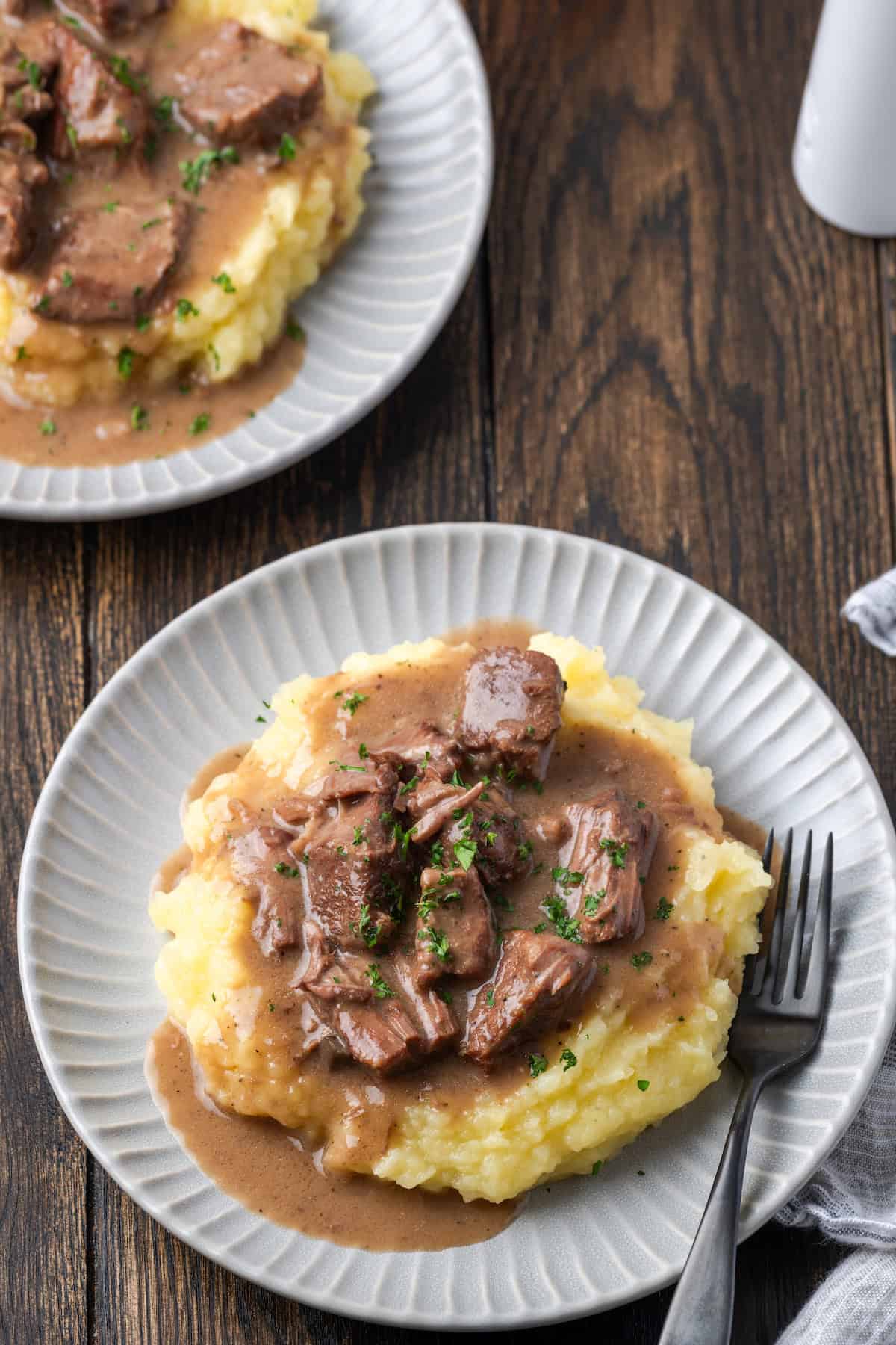 No peeping beef tips and gravy served over mashed potatoes on a white plate next to a fork with a second plate of beef tips in the background.