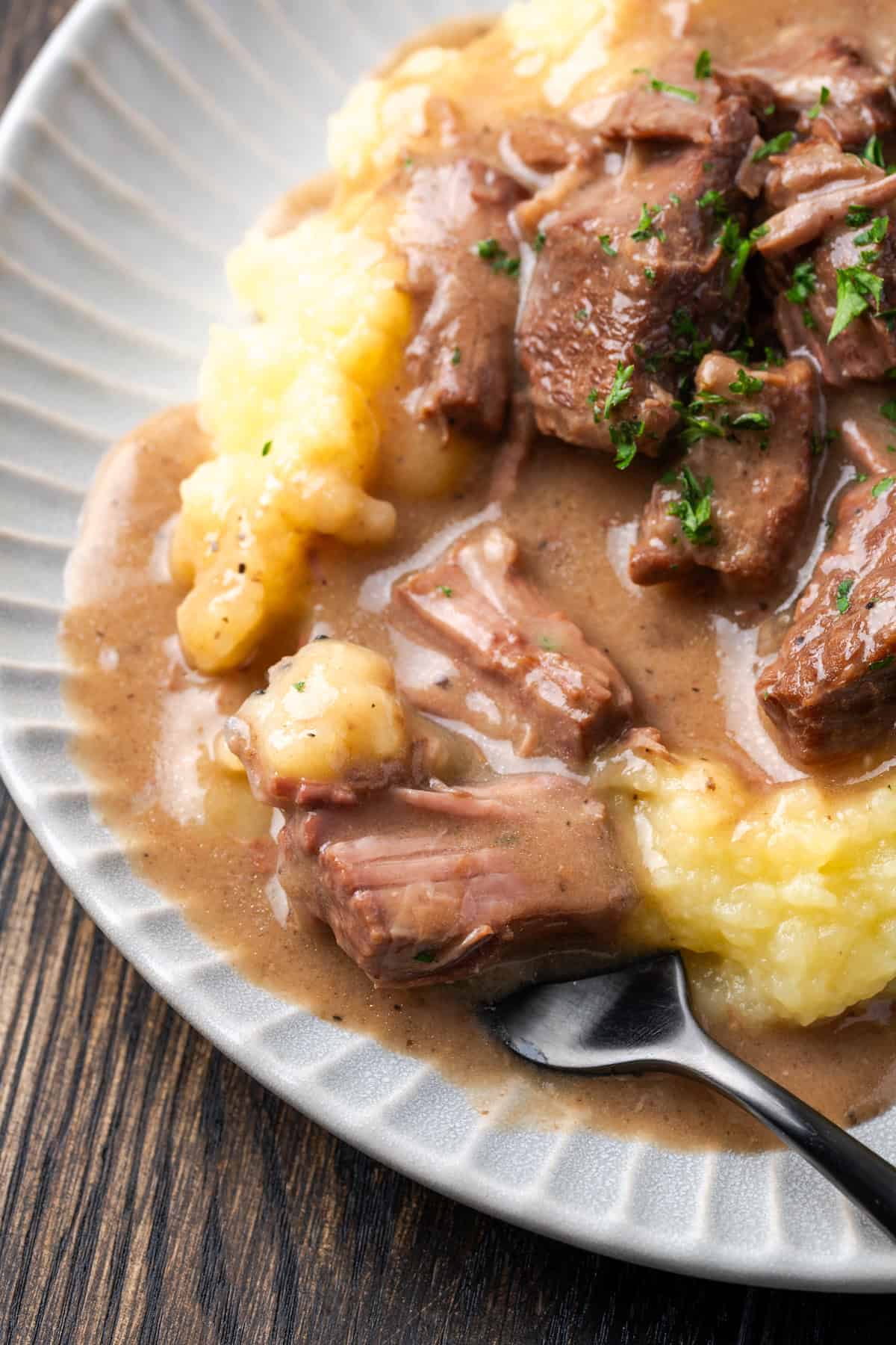 Close up of a fork resting next to mashed potatoes topped with beef tips and gravy on a white plate.