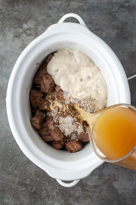 Beef broth is poured over beef tips with cream of mushroom soup and seasonings in a crock pot.