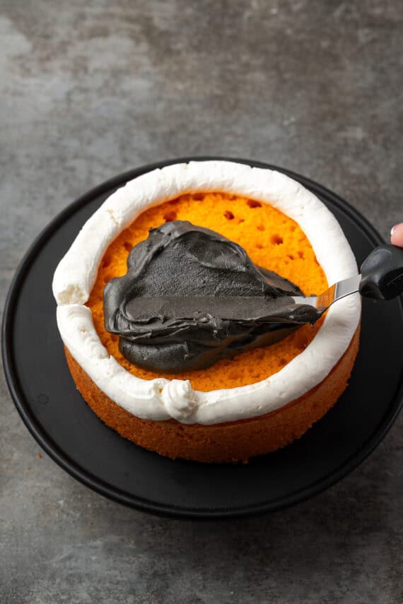 An offset spatula spreading black chocolate frosting over an orange cake layer, inside a piped vanilla frosting border.