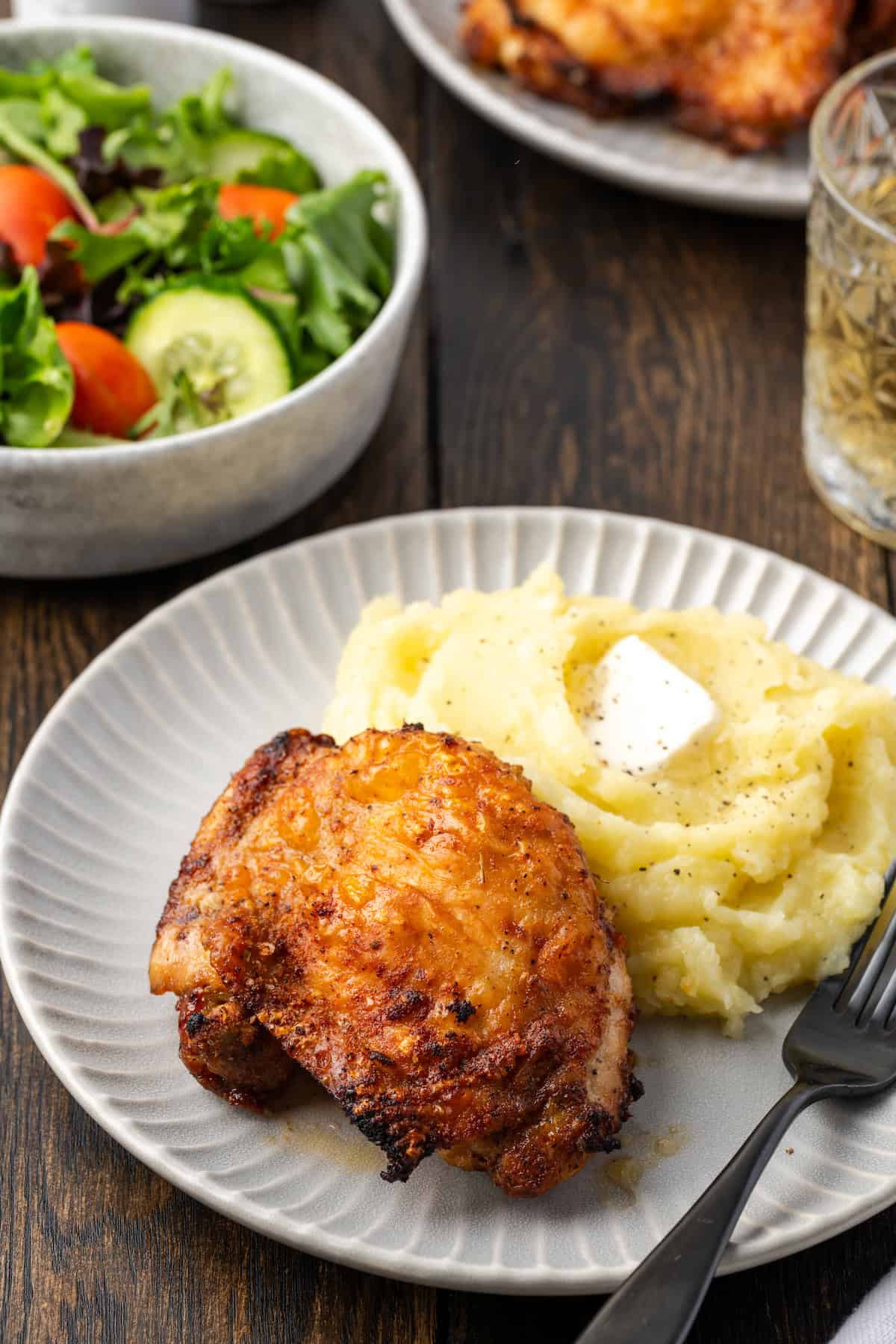 An air fryer chicken thigh served next to mashed potatoes on a plate, with a bowl of salad and a platter of chicken thighs in the background.