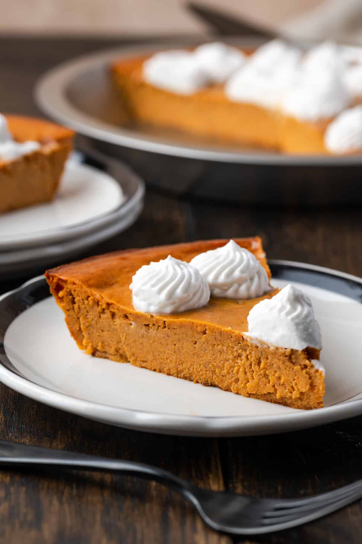 A slice of forkless pumpkin pie with a fork on a plate, the rest of the pie in the background.