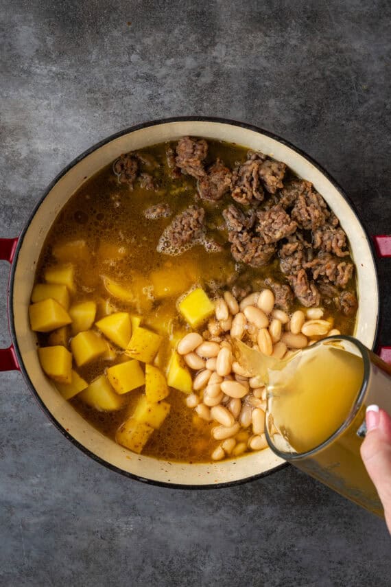 Broth being poured into a pot over top of potatoes, beans, and sausage.