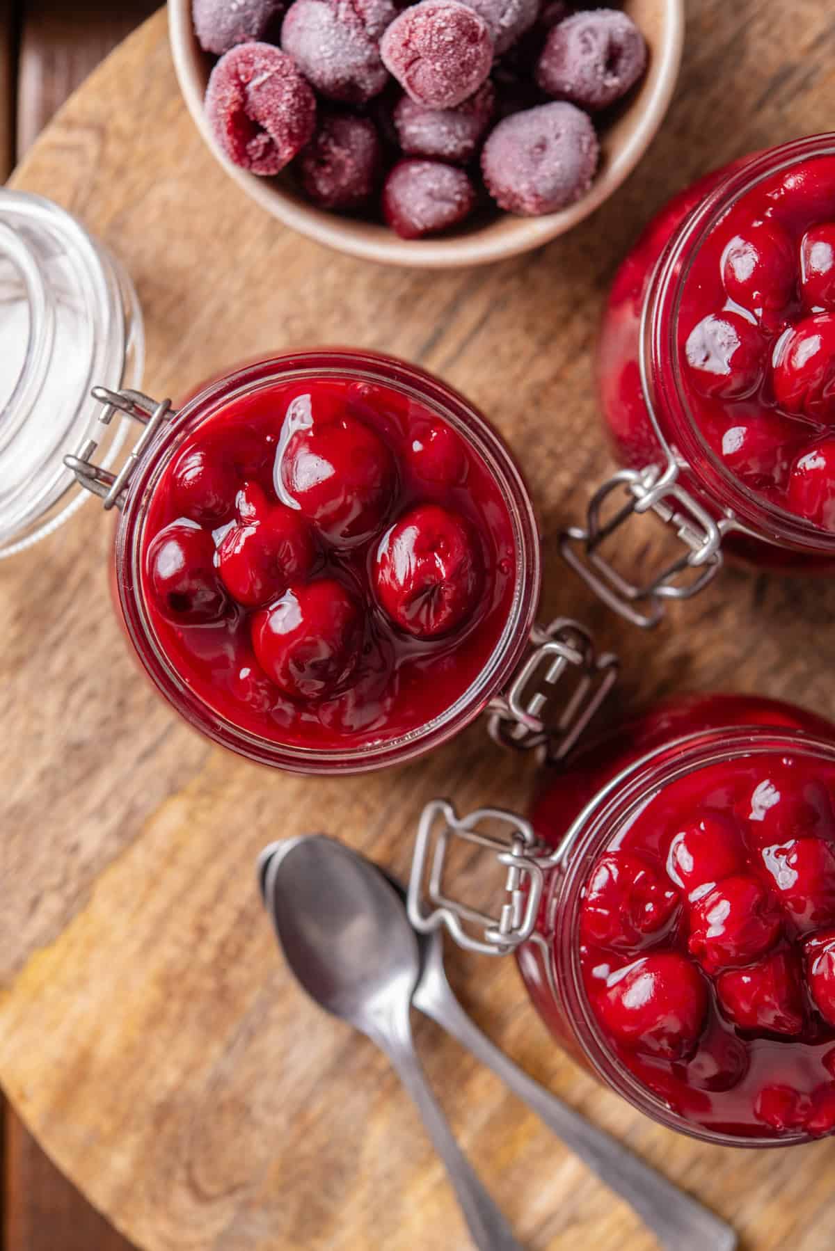 Overhead view of three open jars of cherry pie filling on a round wooden tray, next to a bowl of frozen cherries.