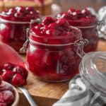 Three open jars of cherry pie filling on a round wooden tray, next to a spoonful of pie filling.