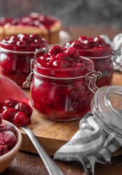 Three open jars of cherry pie filling on a round wooden tray, next to a spoonful of pie filling.