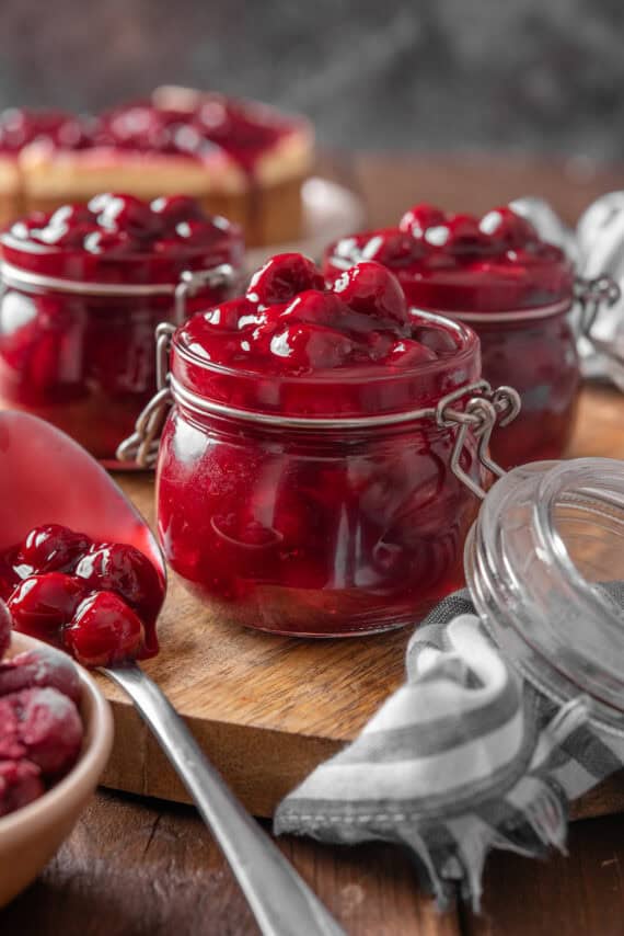 Three open jars of cherry pie filling on a round wooden tray, next to a spoonful of pie filling.