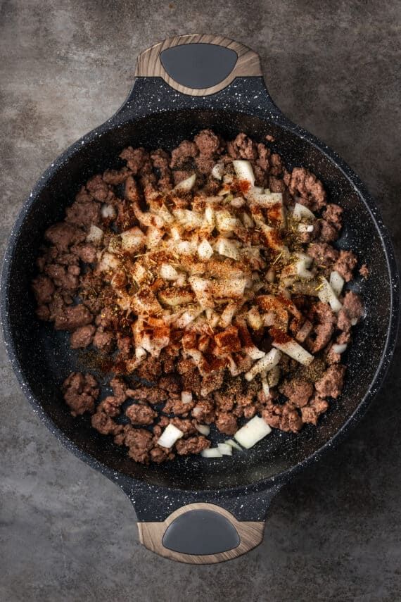 Diced onion and seasonings added to browned ground beef in a skillet.