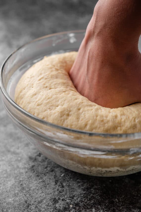 A hand punching down the risen dough in a large glass bowl.