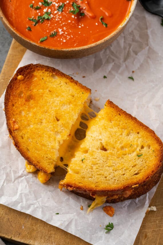 Close up of an air fryer grilled cheese cut in half and slightly pulled apart to reveal the strings of cheese inside, next to a bowl of tomato soup.