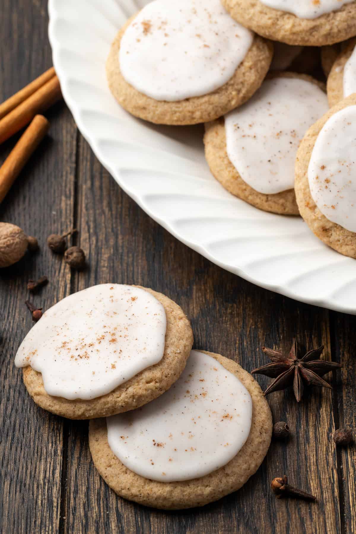 Two chai sugar cookies on a wooden table next to more chai cookies on a white platter.