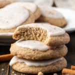 Three chai sugar cookies stacked on a wooden tabletop with a bite missing from the top cookie, and a plate of cookies in the background.