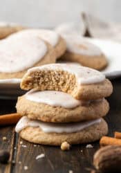 Three chai sugar cookies stacked on a wooden tabletop with a bite missing from the top cookie, and a plate of cookies in the background.