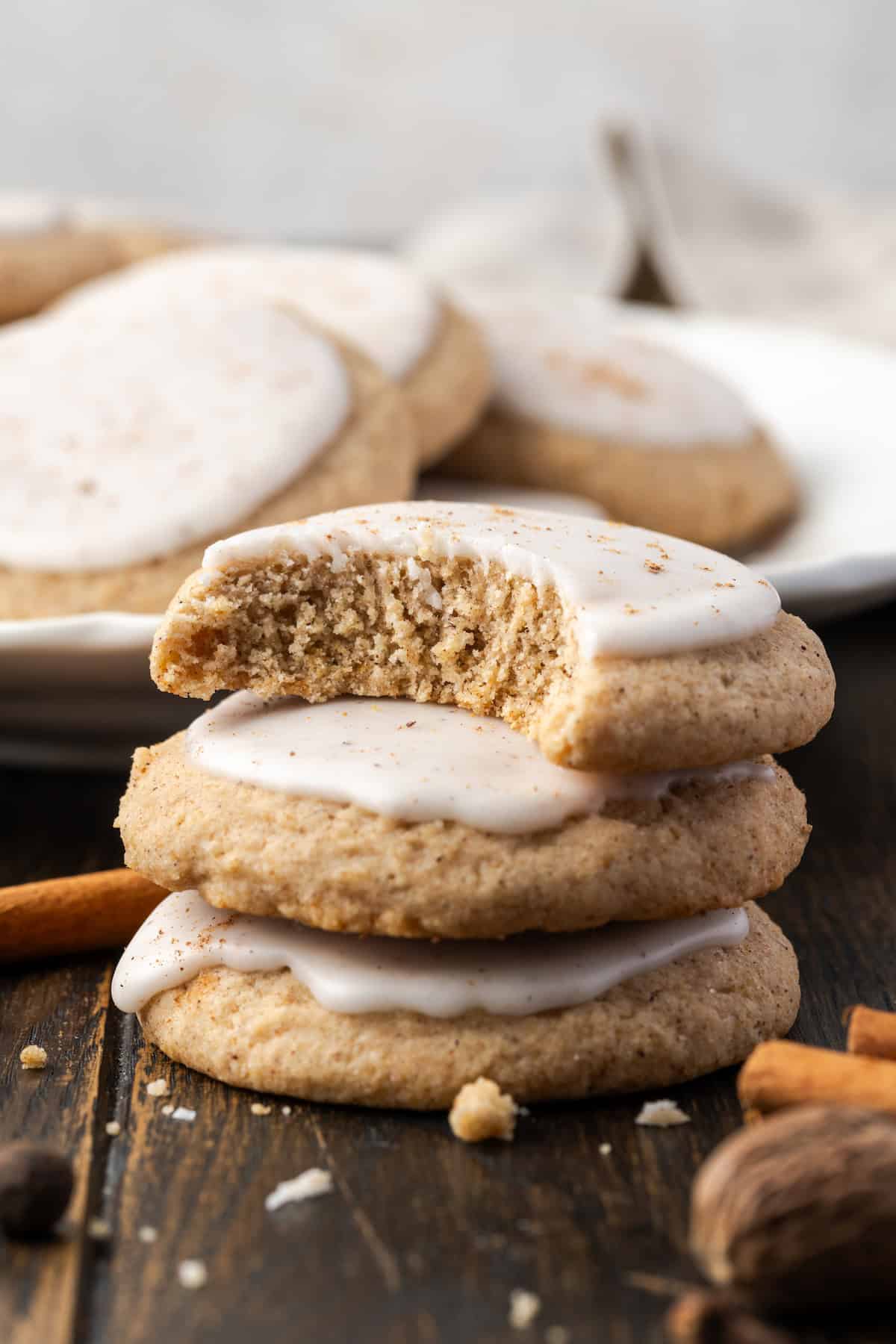 Three chai sugar cookies stacked on a wooden tabletop with a bite missing from the top cookie, and a plate of cookies in the background.