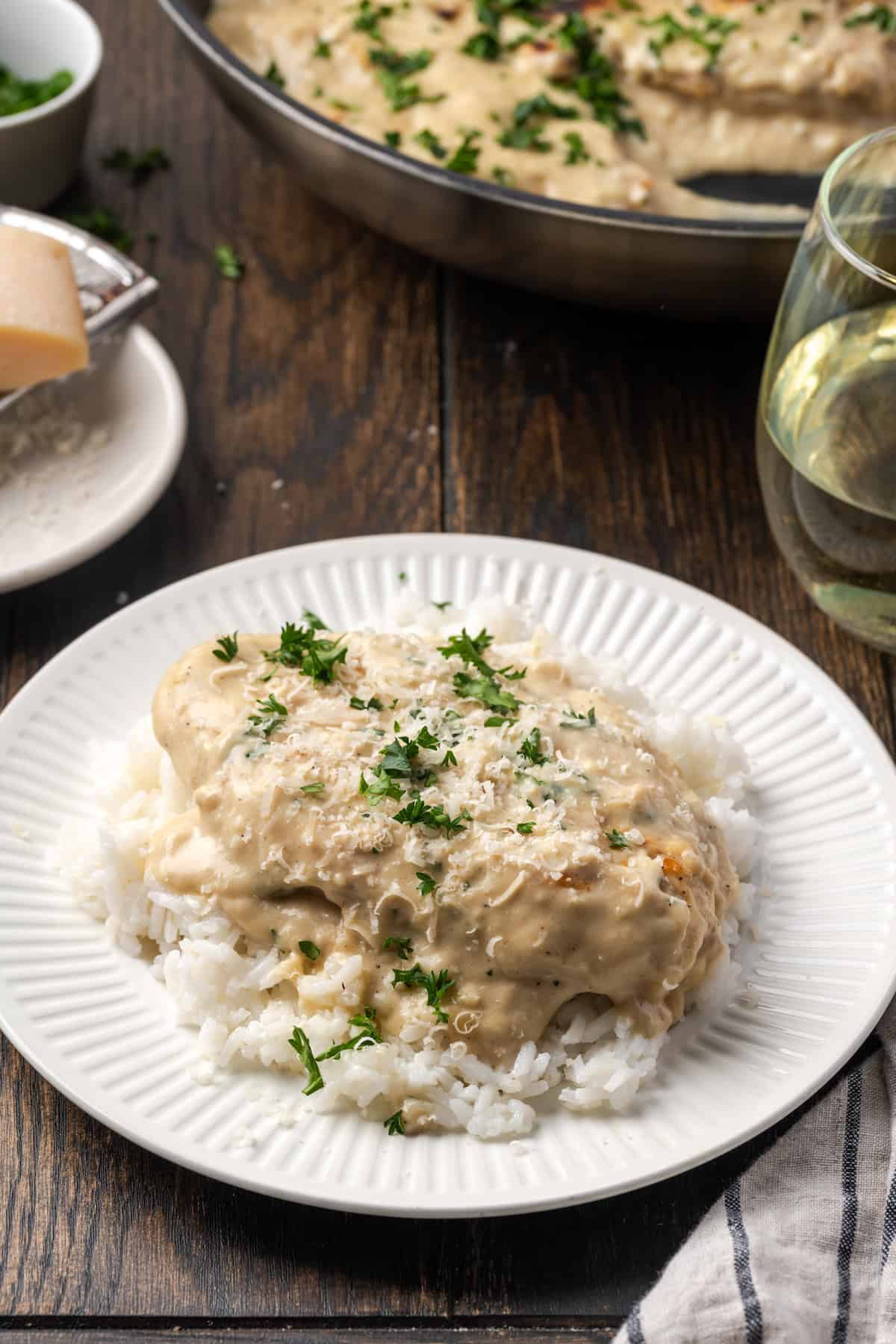 Garlic parmesan chicken served over a bed of white rice on a plate, with more chicken in a skillet in the background.