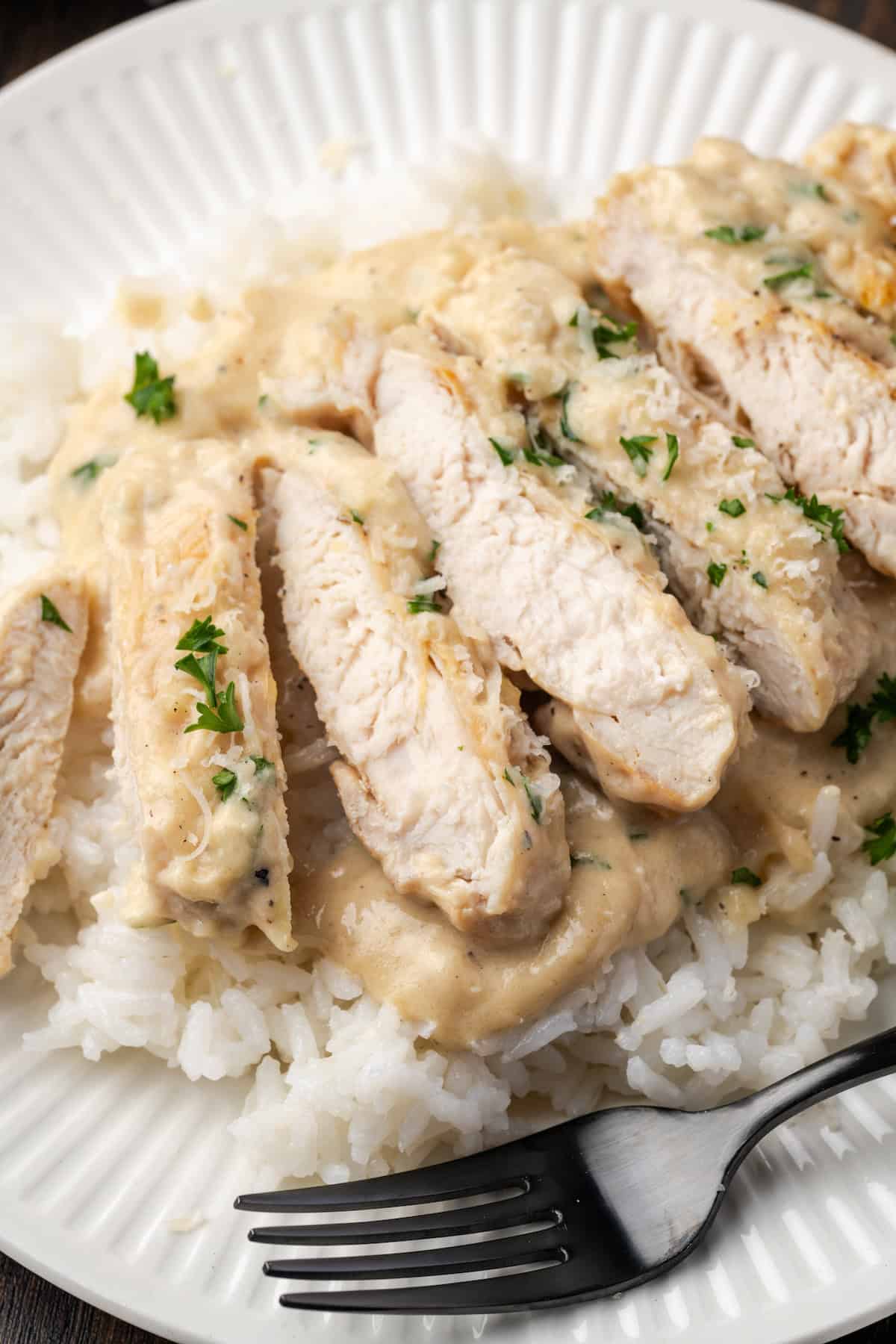 Close up of garlic parmesan chicken breast cut into slices over a bed of white rice on a plate, next to a fork.