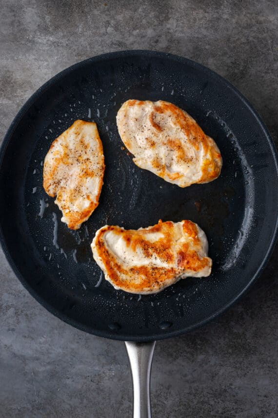 Three chicken breasts searing in a skillet.