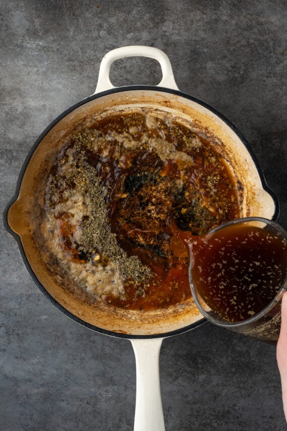 Seasoned beef broth being poured into a roux of flour and butter in a skillet.