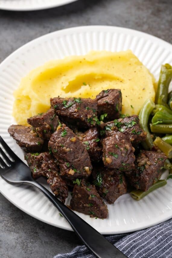 Air fryer steak bites next to a side of mashed potatoes and green beans on a white plate, with a fork.