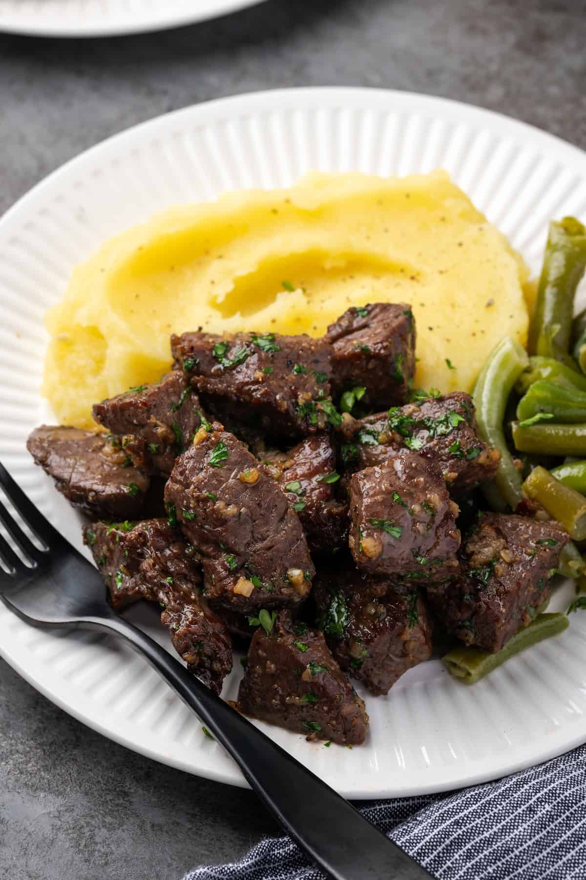 The air fryer steak bite the mashed potatoes and green beans with the fork on a white plate.