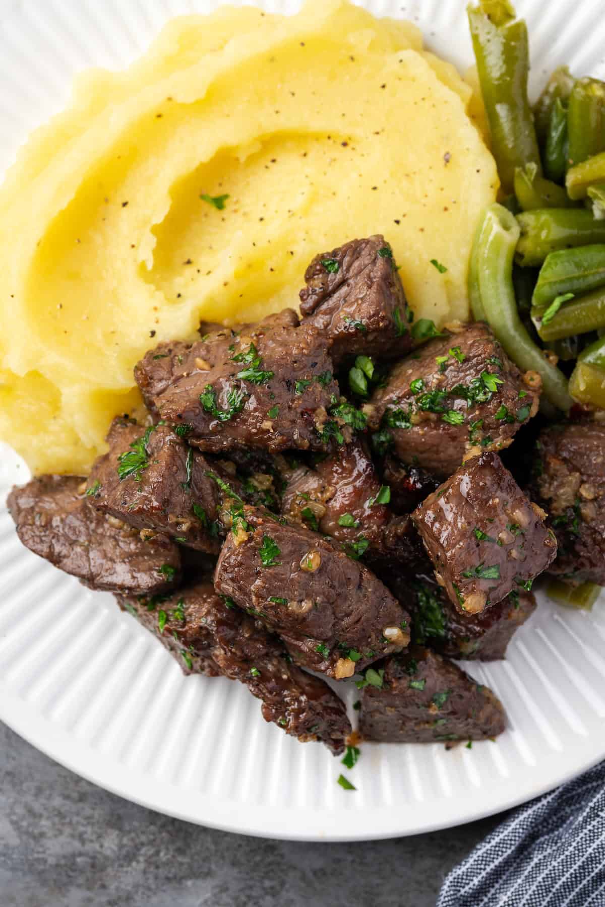 Close up of air fryer steak bites next to a side of mashed potatoes and green beans on a white plate.