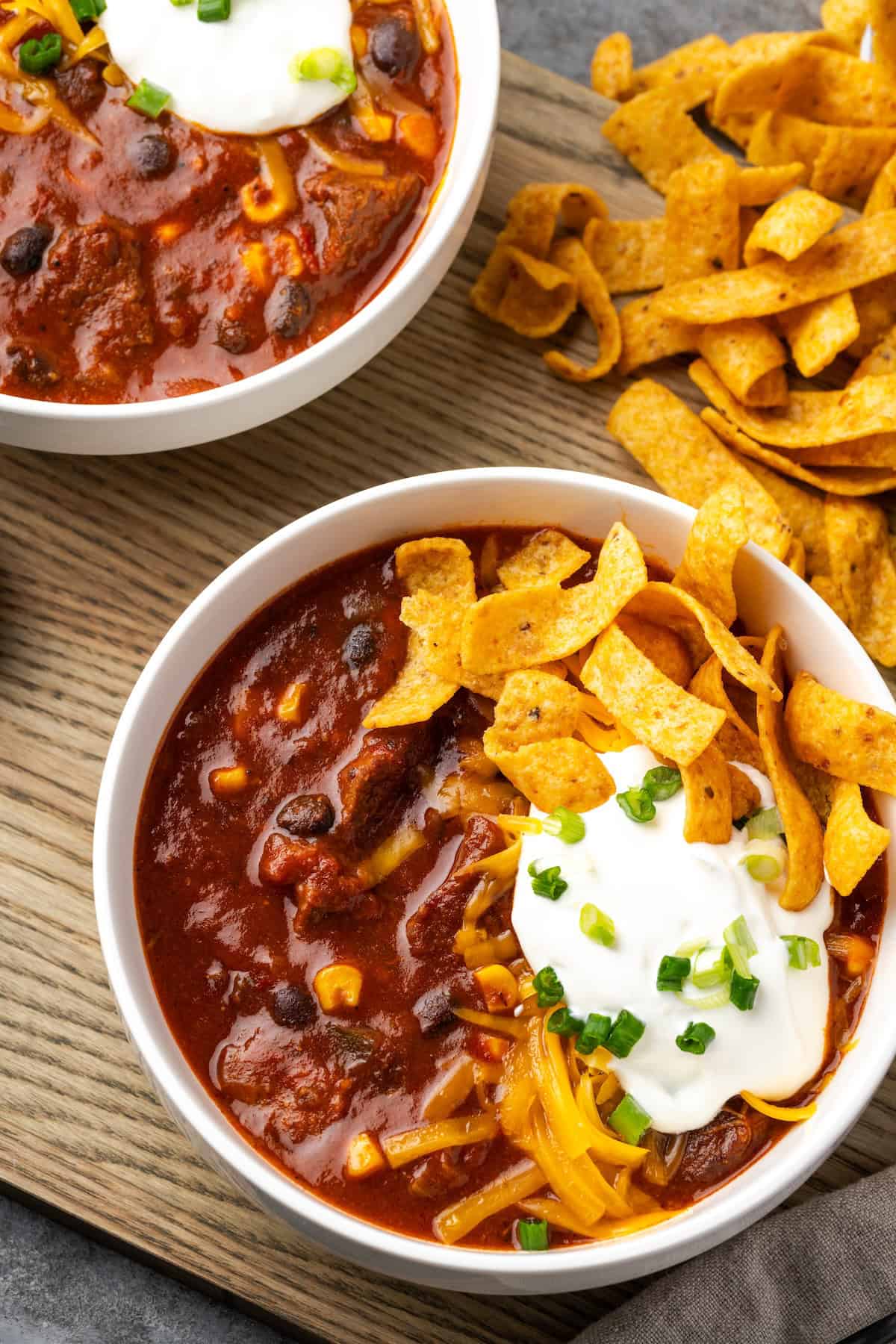 Overhead view of a bowl of chili con carne topped with sour cream and Fritos chips, next to more Fritos and a second bowl of chili.
