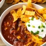 A bowl of chili con carne topped with sour cream and Fritos chips, with more Fritos in the background.