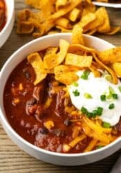A bowl of chili con carne topped with sour cream and Fritos chips, with more Fritos in the background.