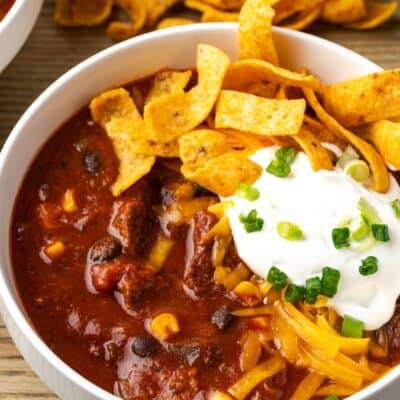A bowl of chili con carne topped with sour cream and Fritos chips, with more Fritos in the background.