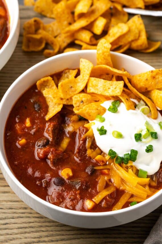 A bowl of chili con carne topped with sour cream and Fritos chips, with more Fritos in the background.