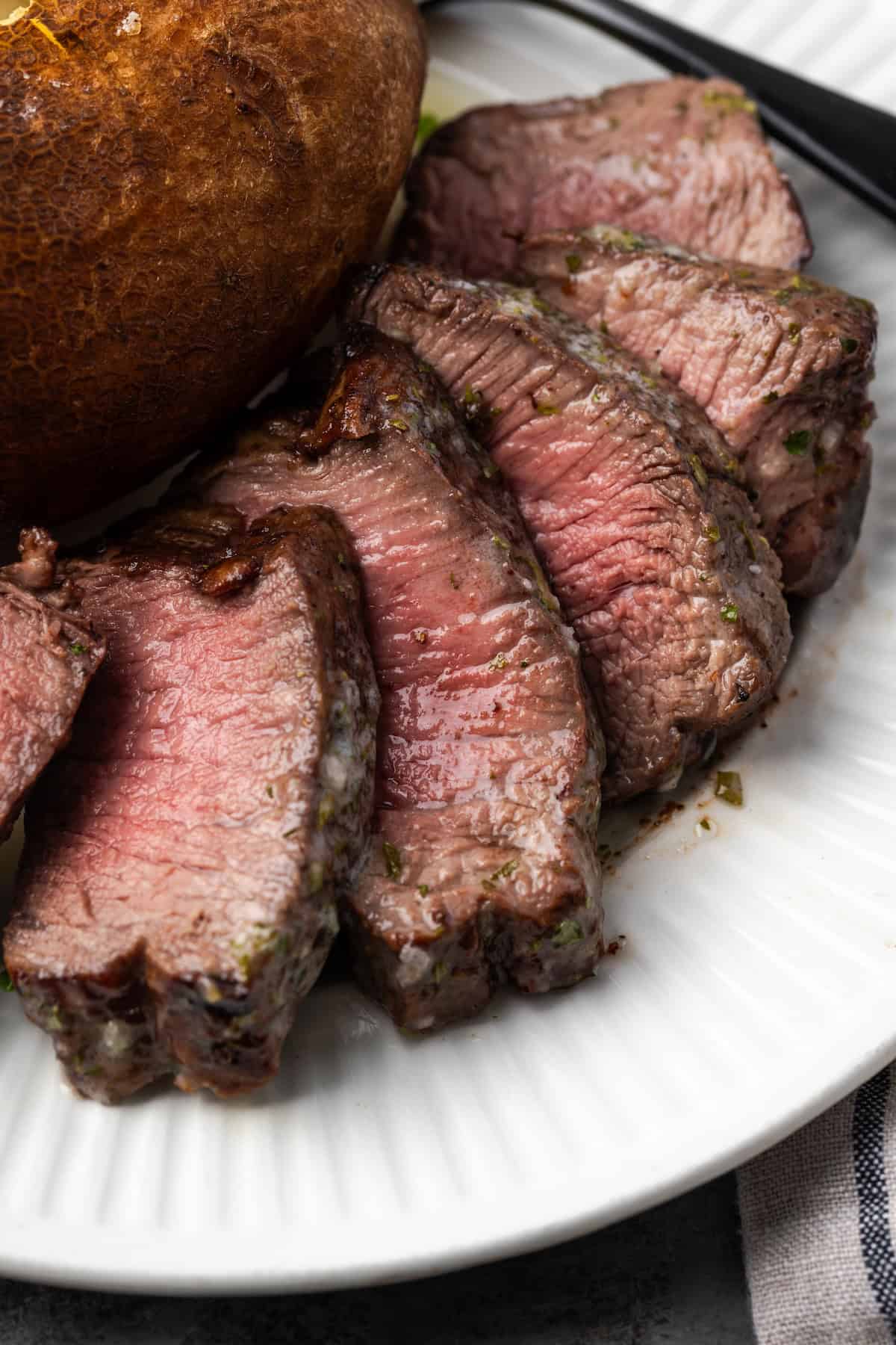 Make juicy, restaurant-quality air fryer filet mignon slathered with homemade garlic compound butter for the ultimate steak dinner! Close up of air fryer filet mignon cut into slices next to a baked potato on a plate.
