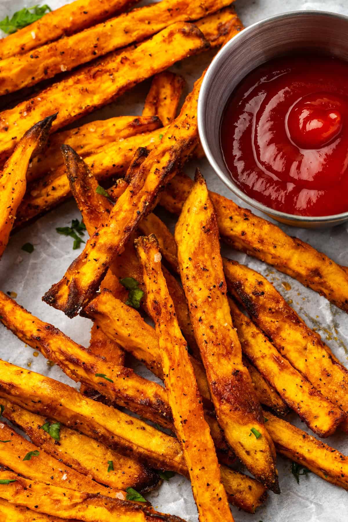 Seasoned with garlic and paprika, crispy air fryer sweet potato fries are tasty and easy to make. It's quicker and lighter than deep frying! Close up overhead view of air fryer sweet potato fries on a parchment-lined plate next to a ramekin of ketchup.