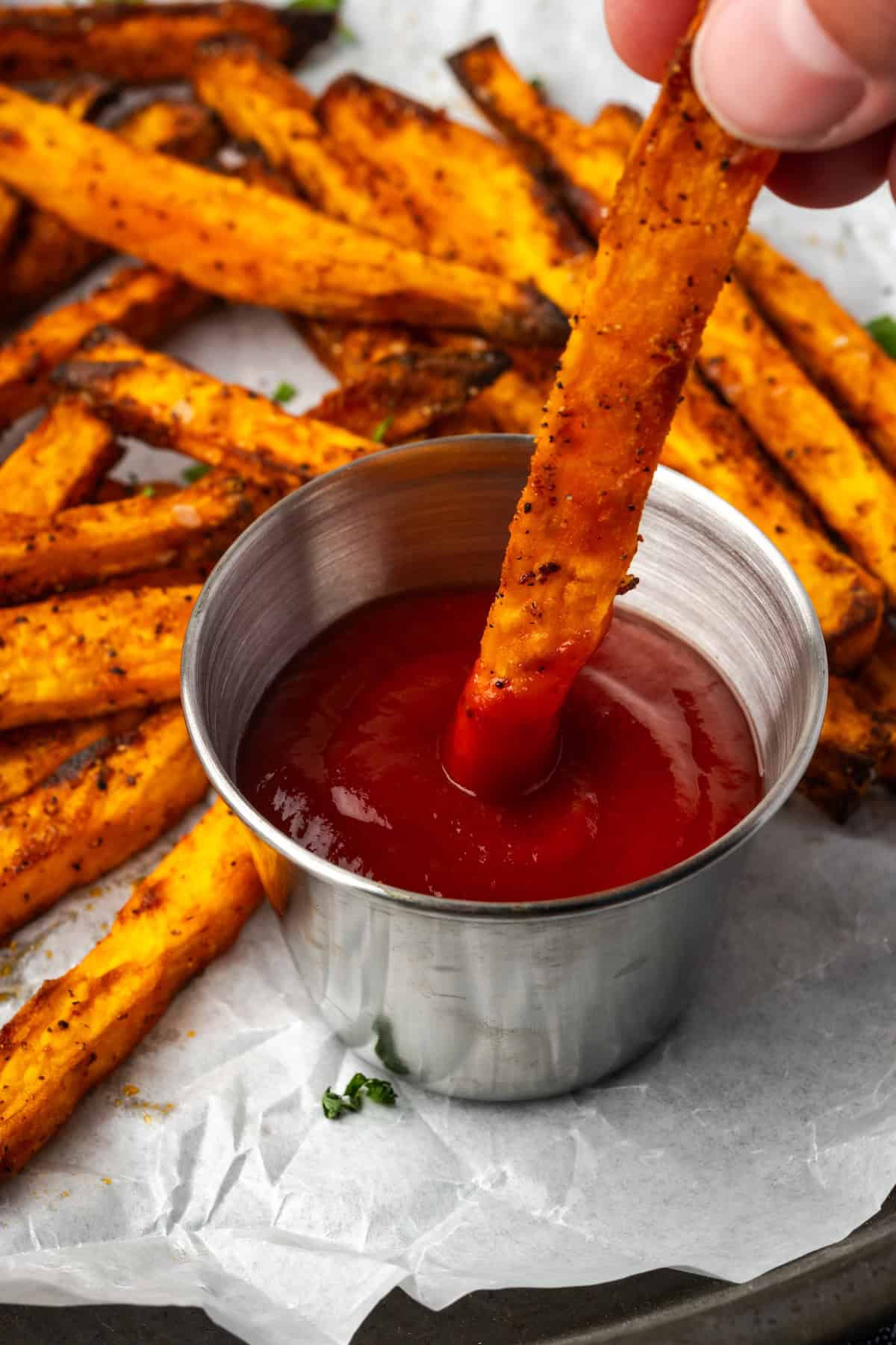 Seasoned with garlic and paprika, crispy air fryer sweet potato fries are tasty and easy to make. It's quicker and lighter than deep frying! A hand dipping a sweet potato fry into a ramekin of ketchup, with more air fryer sweet potato fries in the background.