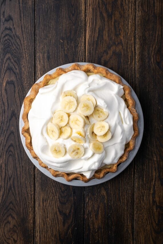 This banana cream pie recipe is a classic dessert with a rich vanilla custard layered with heaps of whipped cream and sweet banana slices. Overhead view of the finished banana cream pie topped with whipped cream and banana slices.