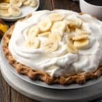 A whole banana cream pie topped with whipped cream and banana slices with a cup of coffee in the background.
