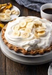 A whole banana cream pie topped with whipped cream and banana slices with a cup of coffee in the background.
