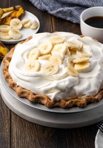 A whole banana cream pie topped with whipped cream and banana slices with a cup of coffee in the background.