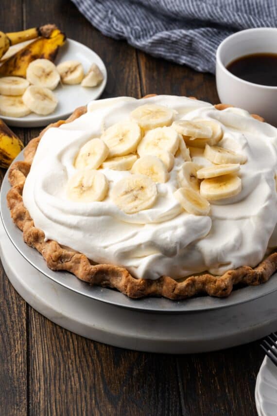 A whole banana cream pie topped with whipped cream and banana slices with a cup of coffee in the background.